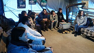9 junge Menschen sitzen in einem Versammlungszelt neben einem Holzofen und hören zu.