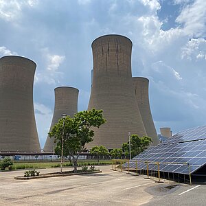 Ein großes Kohlekraftwerk mit vier monumentalen Kühltürmen dominiert das Bild. Im Vordergrund befinden sich Solarzellen auf einer schrägen Konstruktion, umgeben von einem Zaun. Einige Bäume und Büsche sorgen für etwas Grün in der Szene. Der Himmel ist leicht bewölkt.