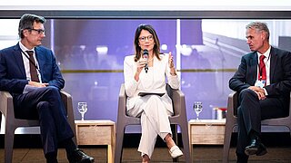 Ana Toni sits on a podium, framed by two men