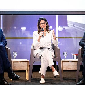 Ana Toni sits on a podium, framed by two men