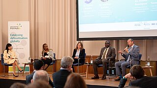 Ein Podium mit fünf Teilnehmenden sitzt auf der Bühne während einer Konferenzveranstaltung, die vom International PtX Hub organisiert wird. Im Hintergrund steht ein Banner mit der Aufschrift „Katalysierung der Defossilisierung weltweit“. Ein Mann ganz rechts spricht ins Mikrofon, während die anderen aufmerksam zuhören.