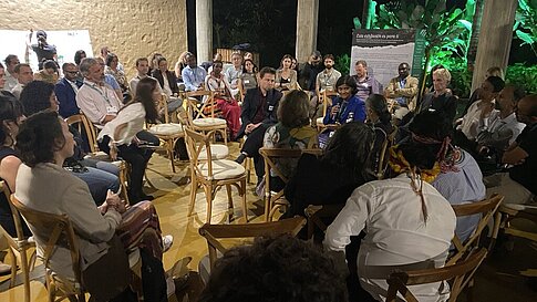 An engaged audience sits in a circular setup, attentively listening to a speaker in a blue jacket holding a microphone. The room has wooden decor, and greenery is visible in the background, giving the space a natural ambiance.