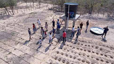 Aerial view of a group of people gathered around a water tank structure in a dry, rural area. Some individuals are examining the tank while others stand nearby, observing or talking. The ground is arranged with rows of shallow, freshly dug planting beds, and irrigation lines are visible. Sparse trees and brush surround the scene, and a large metal dish lies on the ground to the right.