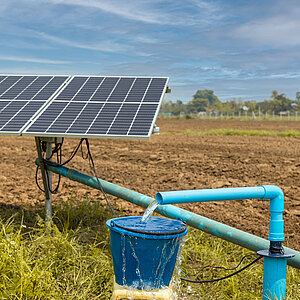 Solarbetriebene Wasserpumpe auf einem Feld: Eine blaue Wasserpumpe pumpt Wasser in einen Eimer, angetrieben durch Solarmodule im Hintergrund. Das Feld ist trocken und von grünem Gras umrandet.t