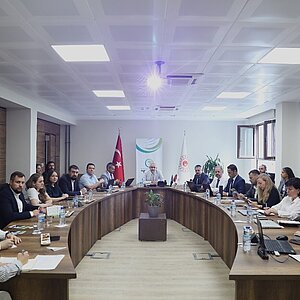 A conference room with officials and delegates seated around a large, U-shaped wooden table. The meeting appears to be formal, with participants using laptops and writing notes. The Turkish flag and an organizational banner are visible behind the central figure leading the discussion.