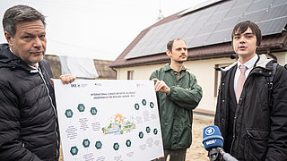 [Translate to English:] Federal Minister for Economic Affairs and Climate Action Robert Habeck holds an IKI chart with another man. To the right stands a young man who is being interviewed.