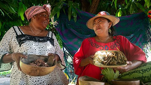 Two women in traditional clothing smile at each other, holding bowls of food under a tree.