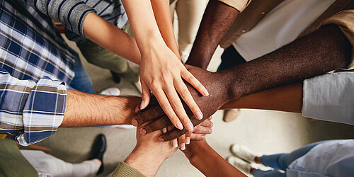 People with different skin colours stand in a circle and hold their hands over each other in the middle