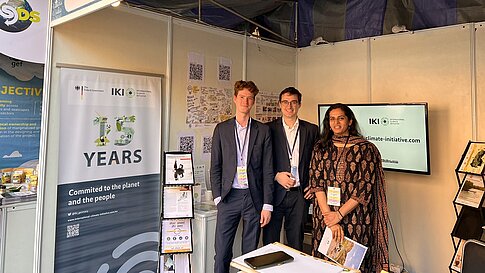 2 Männer und eine Frau stehen in einem Messestand. Auf der linken Seite ist ein Roll-up mit dem IKI-Jubiläumslogo zu sehen. 