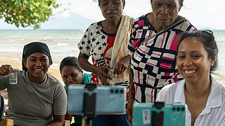 5 black women sitting and standing on the beach. Smartphones in selfie sticks can be seen in the foreground, recording a video of the people. 