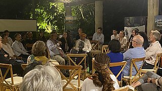 A group of about 20 people are seated in a circle under a roofed outdoor area, surrounded by lush greenery. The attendees are engaged in a discussion, with one man, wearing traditional clothing and a feathered headdress, speaking into a microphone. Banners in the background suggest an event focused on environmental or indigenous topics. The atmosphere is informal and collaborative.