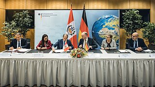 Elmer Schialer, Fey Silva Vidal, Wilbert Rozas Beltrán, Niels Annen, Anja Hajduk and Norbert Gorißen sit at a long table and sign documents.