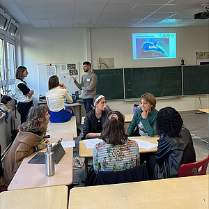 Ein Klassenzimmer mit einer Gruppe von Menschen, die in eine Diskussion vertieft sind. Sechs Teilnehmer sitzen um zwei Tische und besprechen Materialien, die vor ihnen liegen. Im Hintergrund stehen zwei Personen in der Nähe einer Tafel, auf der eine Karte mit der Aufschrift „Community Map of Bedo“ projiziert ist. Eine Person scheint gestikulierend etwas zu erklären oder zu präsentieren. Der Raum hat große Fenster auf der linken Seite, die natürliches Licht hereinlassen, und die Atmosphäre wirkt kollaborativ und konzentriert.t