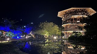 A large, multi-story wooden building with a hexagonal shape is brightly lit at night, its structure reflected in a pond in the foreground. The surrounding area includes trees illuminated in blue and white lights, while a few people can be seen walking near the building. The sky is clear and dark, creating a peaceful atmosphere.