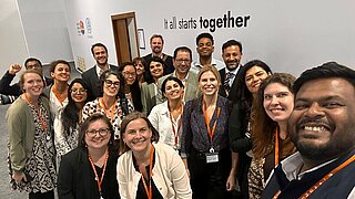 Eine Gruppe von rund zwanzig Menschen posiert lächelnd für ein Gruppenfoto bei einer Networking-Veranstaltung. Sie tragen alle orangefarbene Namensschilder. Im Hintergrund ist eine weiße Wand mit dem Schriftzug „It all starts together“ sichtbar, sowie einige Logos auf der linken Seite.