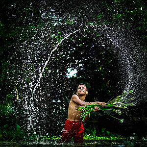 A man with his upper body exposed is standing in a river. He is waving a bundle of branches in both hands. You can see a circle of water drops in the air coming from the branches.